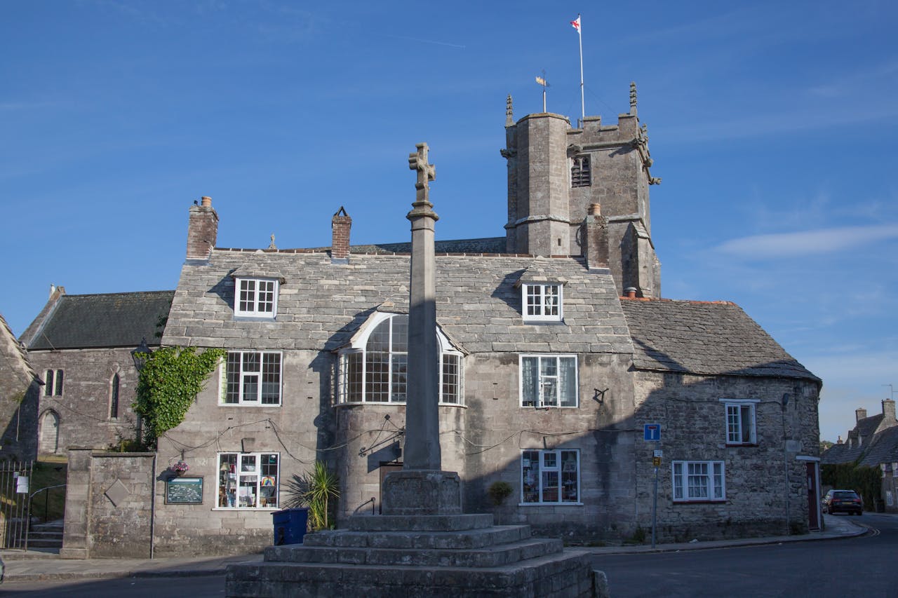 Stone building with a medieval cross monument in Corfe Village, England, under a clear blue sky.